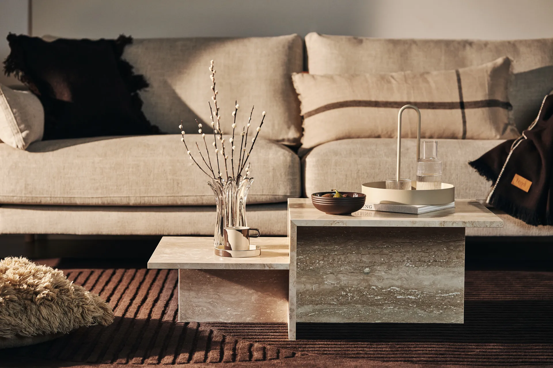 Nivådelat soffbord i beige sten framför en neutral soffa med dekorativa tillbehör.
