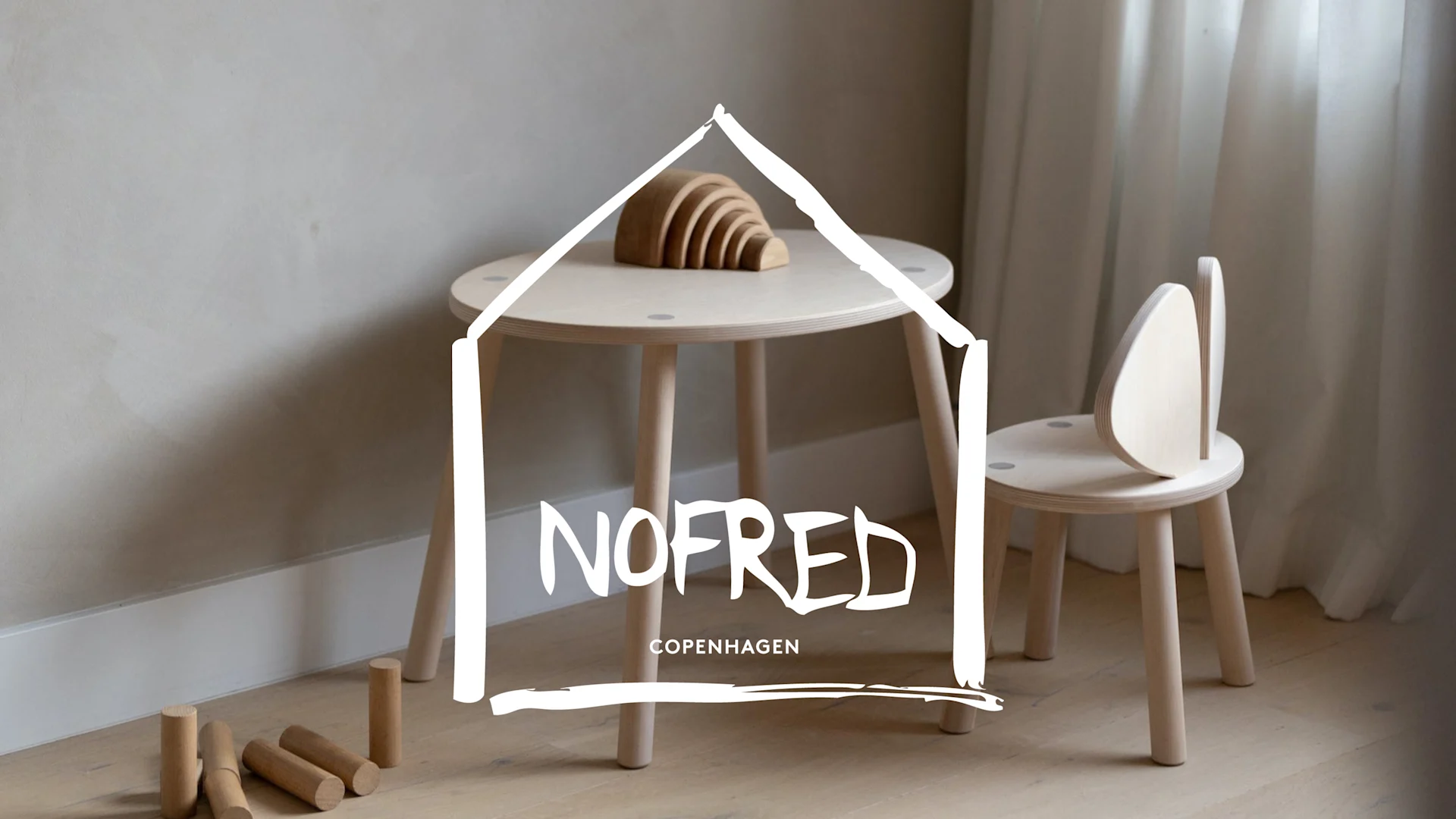 Ett ljust rum med ett runt träbord och trästol, träleksaker och NOFRED COPENHAGEN-logotypen.