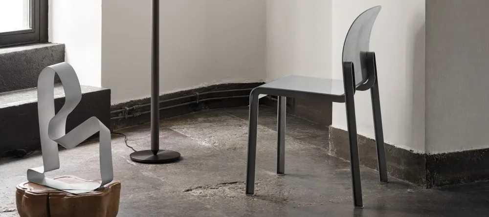 En modern grå stol, en bandliknande skulptur på piedestal och en golvlampa i ett rum.