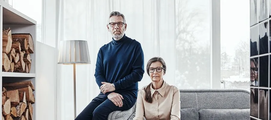 Två äldre personer sitter i ett ljust vardagsrum. Mannen har blå tröja, kvinnan ljusbrun skjorta. Ved och lampa syns.