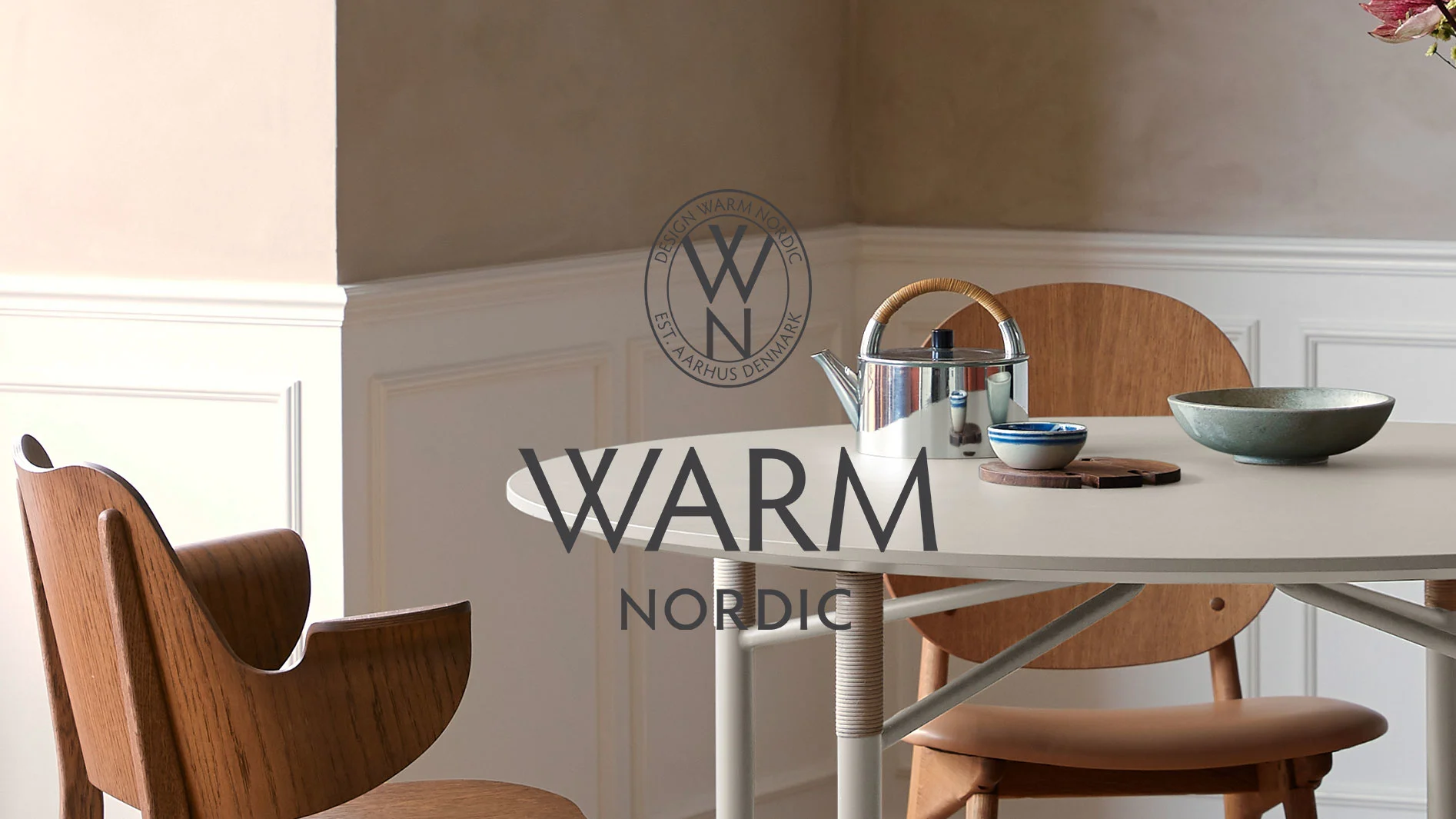 Ett ljust, runt matbord med en silverkanna och keramikskålar, omgivet av trästolar i ett modernt rum med 'Warm Nordic'-logotyp.