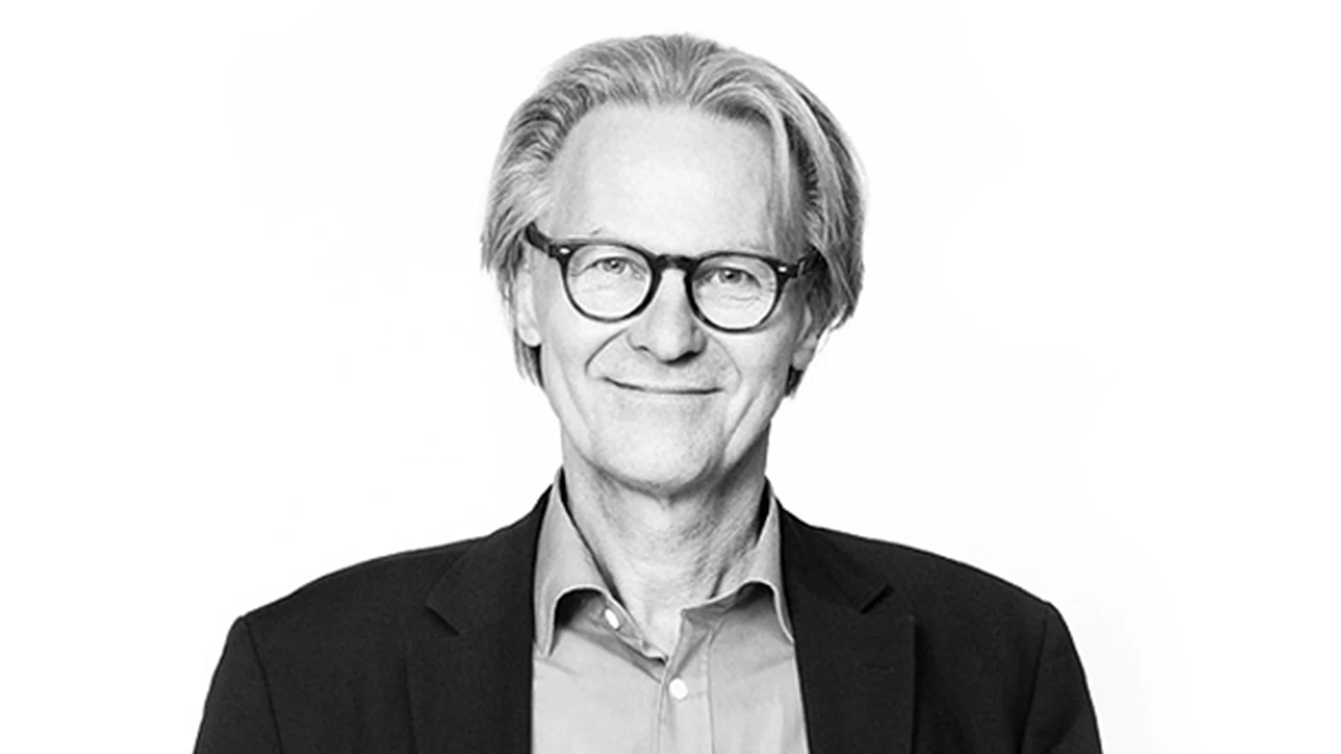Svartvitt porträtt av en leende äldre man med glasögon och grått, tillbakakammat hår, klädd i skjorta och kavaj.