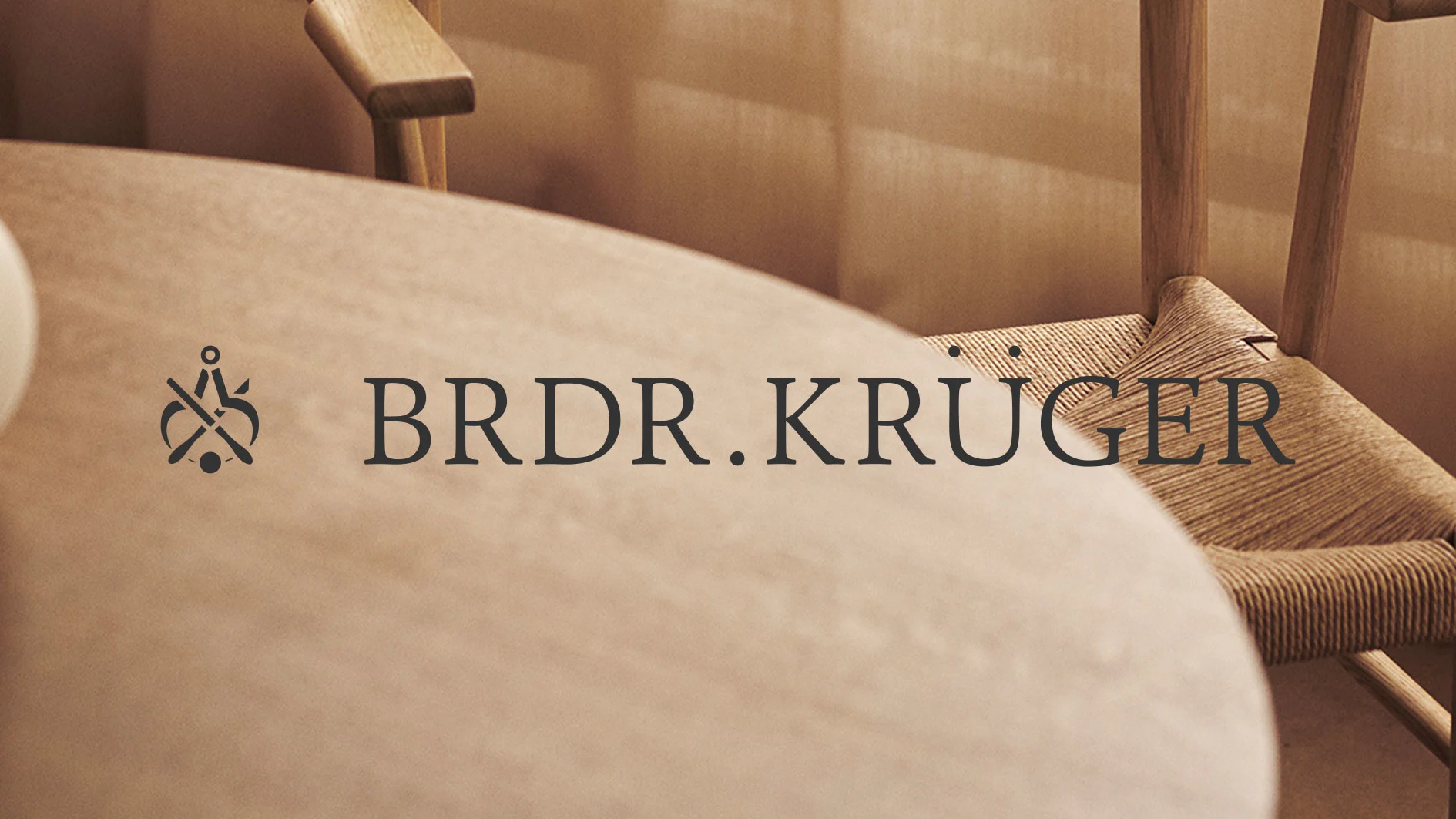 BRDR. KRÜGER-logotyp på en ljus träbordsskiva, med en flätad trästol i bakgrunden.