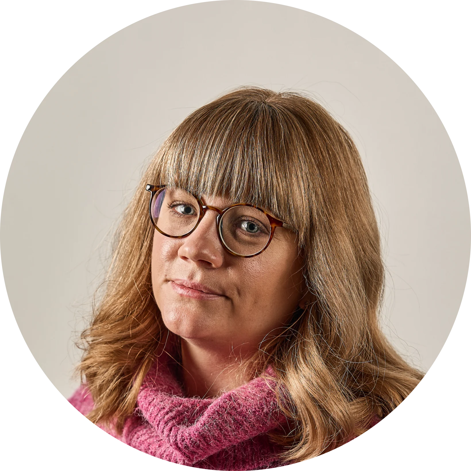 En kvinna med långt blont hår, lugg och glasögon, iklädd en rosa tröja, tittar framåt.