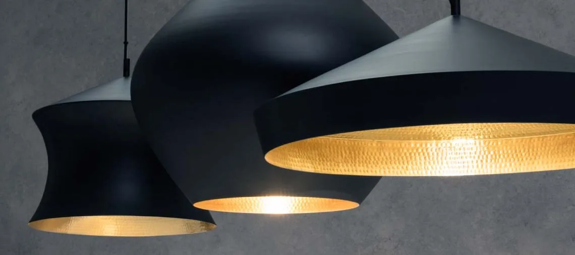 Tre svarta taklampor med hamrade, guldfärgade insidor. De har olika, elegant design och lyser varmt mot en mörk bakgrund.