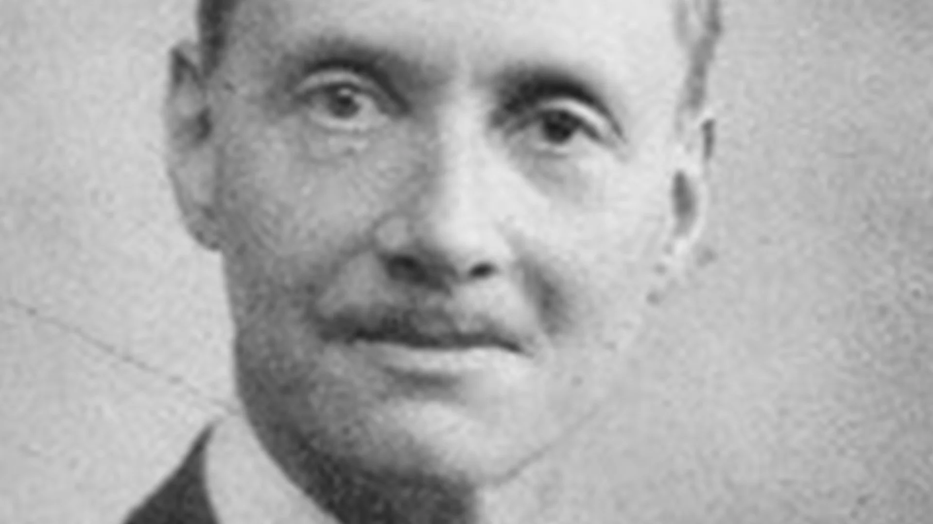 Svartvitt närbild av en man med mustasch som tittar rakt fram.