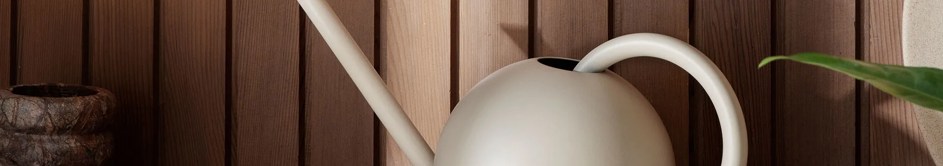 Modern ljusbeige vattenkanna med lång pip och böjt handtag mot en träpanelvägg.