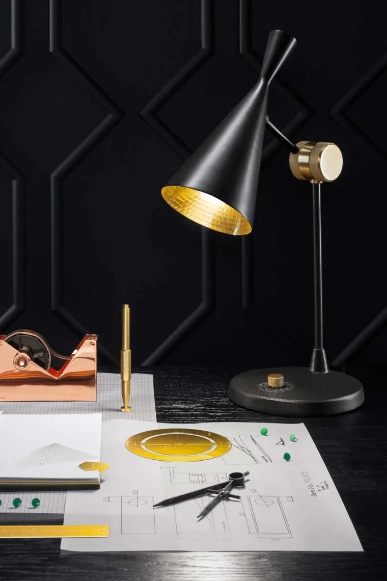 En svart och guld bordslampa på ett mörkt skrivbord med ritverktyg, anteckningsblock och ritpapper.