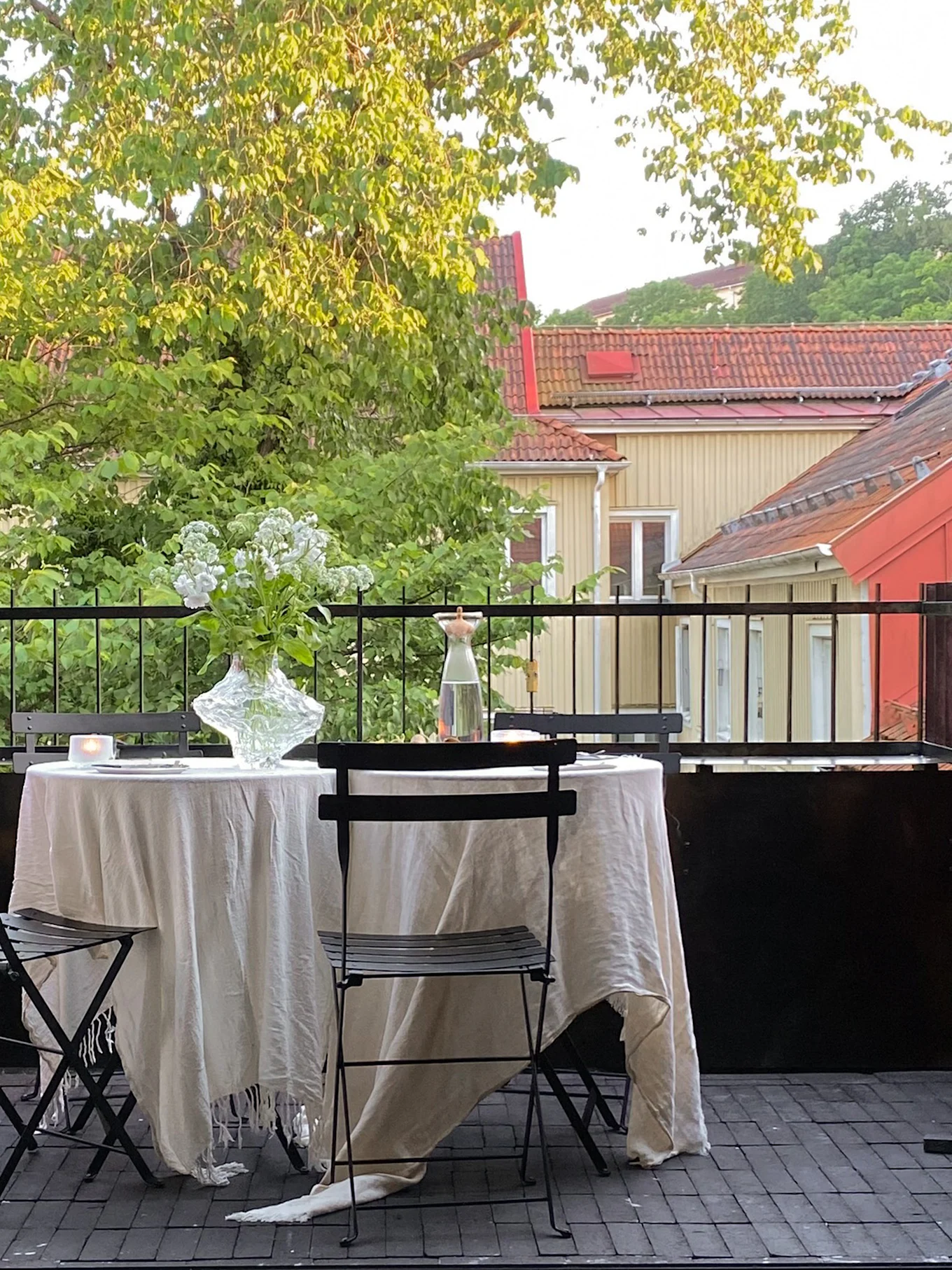 En mysig balkongmatsal med dukat bord, vita blommor och levande ljus, omgiven av gröna träd och hus.