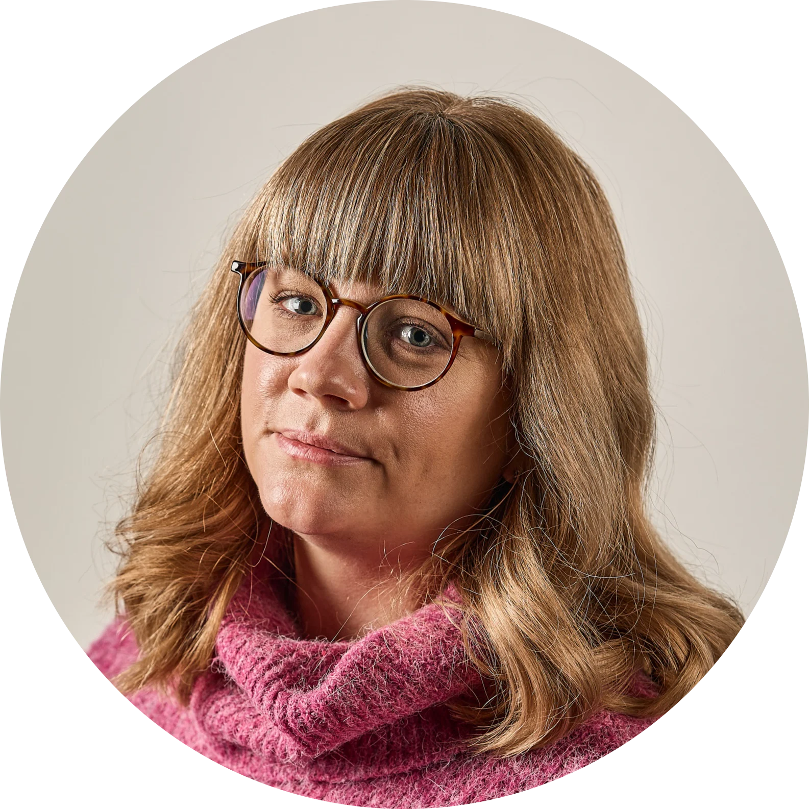 Kvinna med blont hår, lugg, glasögon och rosa tröja, tittar rakt fram.