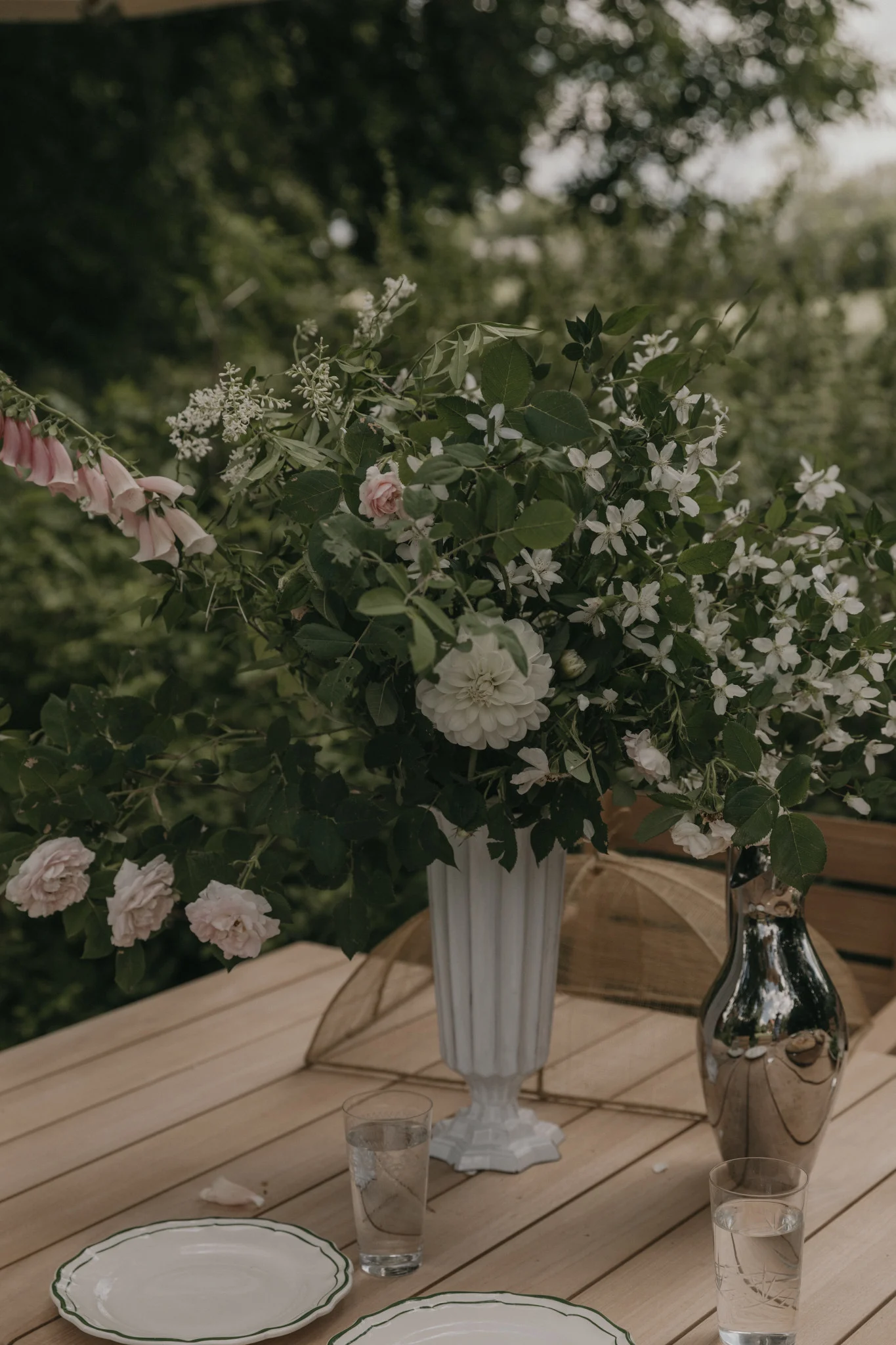 Ett stort blomsterarrangemang med rosa fingerborgsblomma, ljusrosa rosor och vita blommor i en vas på ett träbord.