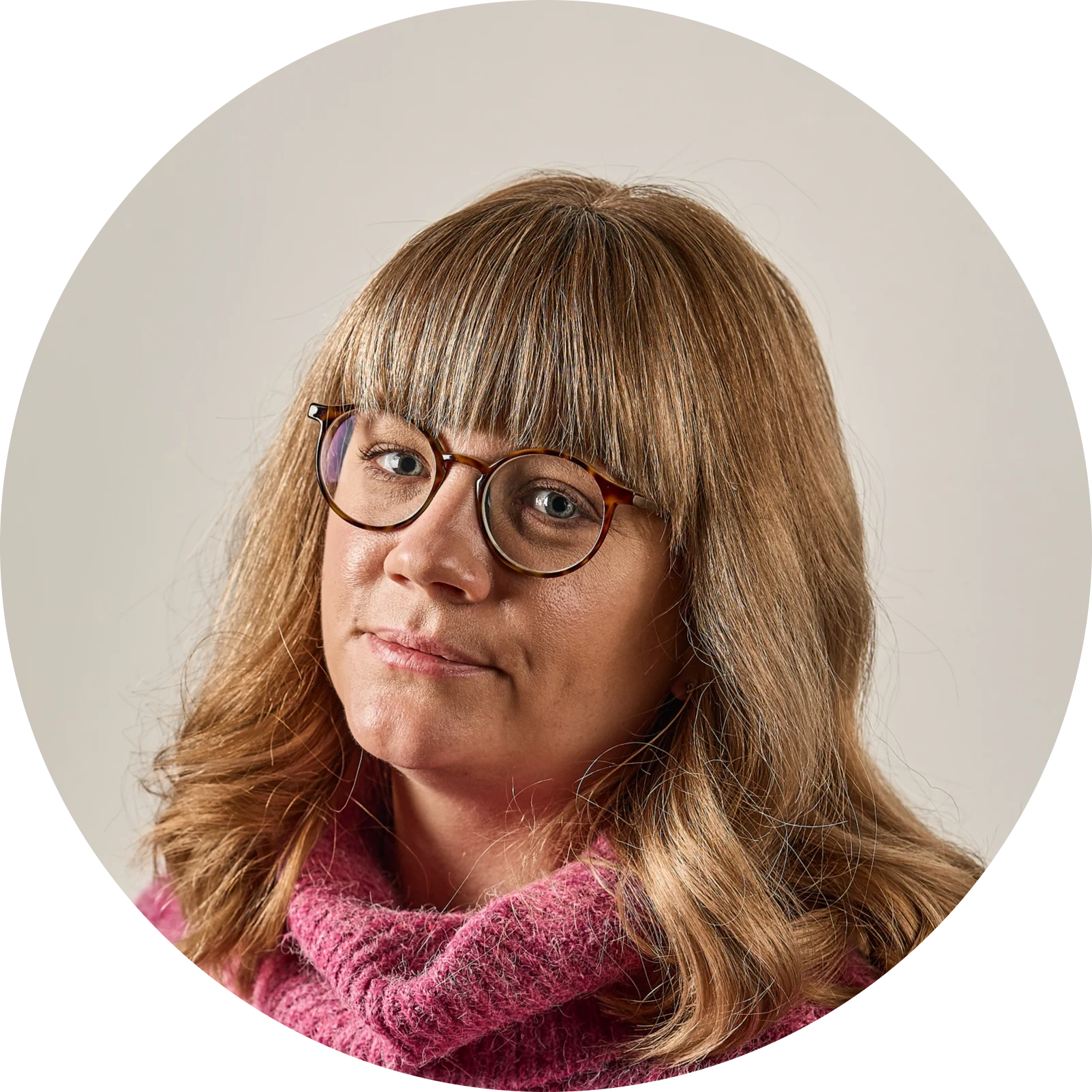 Kvinna med blont hår, lugg, sköldpaddsmönstrade glasögon och rosa tröja, tittar in i kameran.
