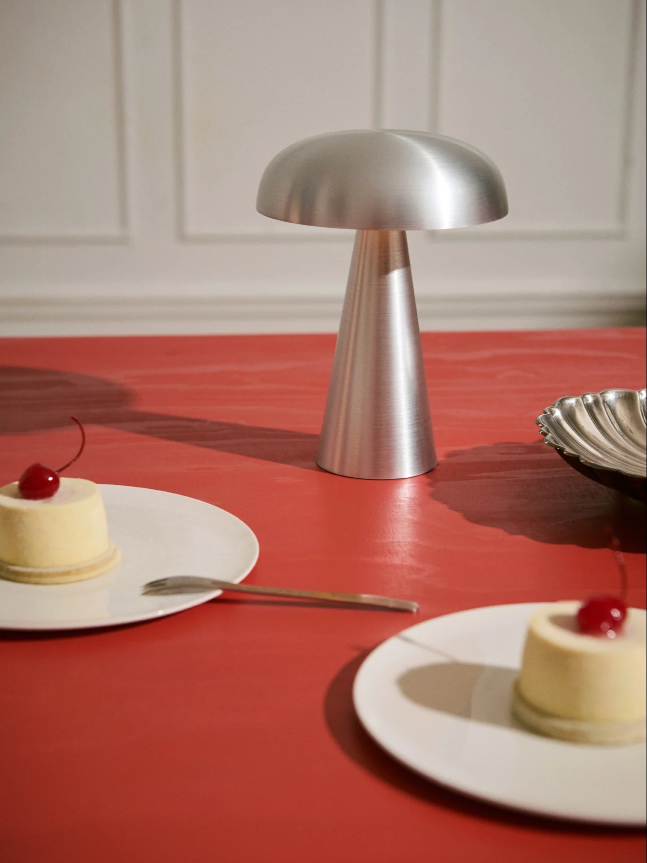 Borstad silverlampa med svampformad topp, konisk bas på rött bord. Två desserter med körsbär.