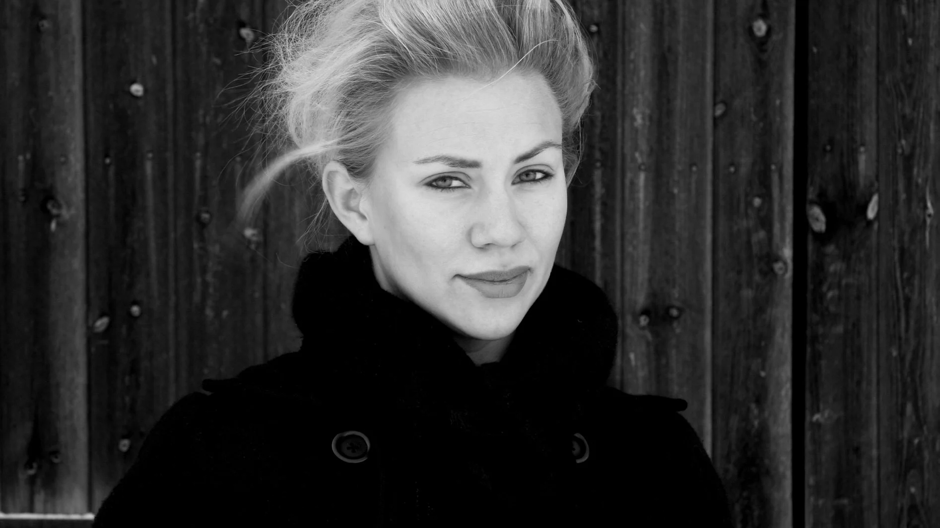 Svartvitt porträtt av en blond kvinna i mörk kappa framför en trävägg.