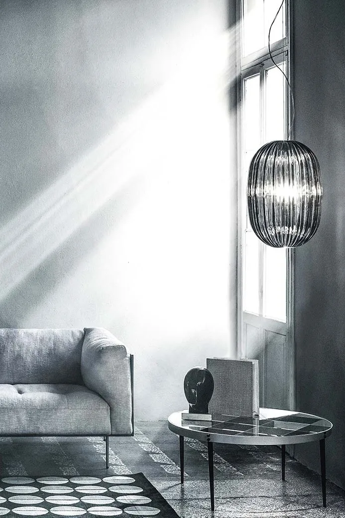 Svartvit bild av ett modernt vardagsrum med en ljus soffa, en räfflad taklampa vid ett soligt fönster och ett runt soffbord med mönstrad skiva.