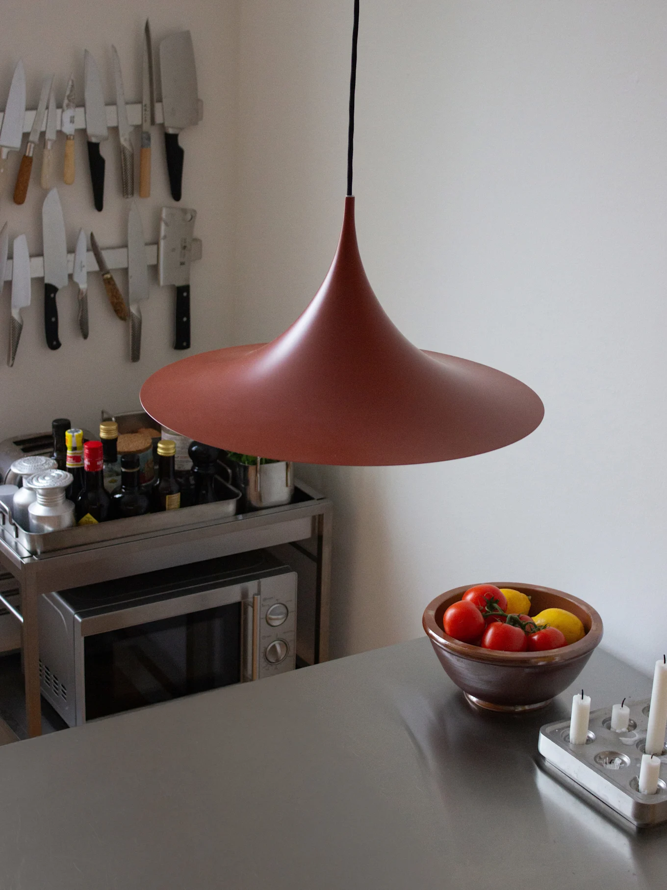 Modernt kök med en röd, trattformad taklampa ovanför en bänkskiva i rostfritt stål, med en skål tomater och citroner.