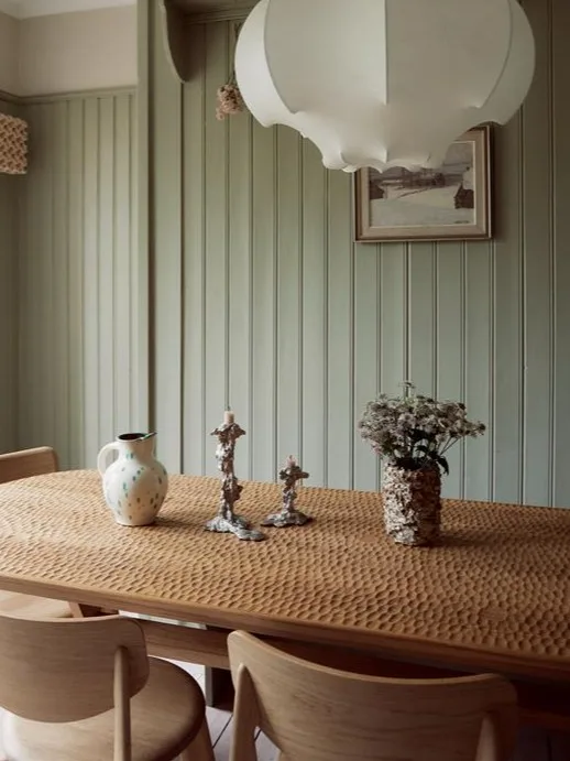 Matbord i trä med bikake-textur, vit kanna, ornamenterade ljusstakar, blommor i vas, gröna panelväggar.