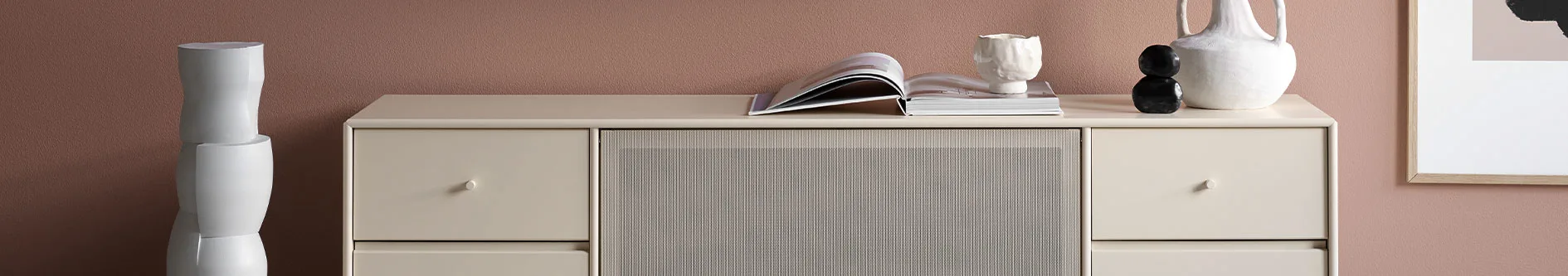 Beige TV-bänk med lådor och en ribbad front, toppad med keramik och en öppen bok, mot en rostbrun vägg.