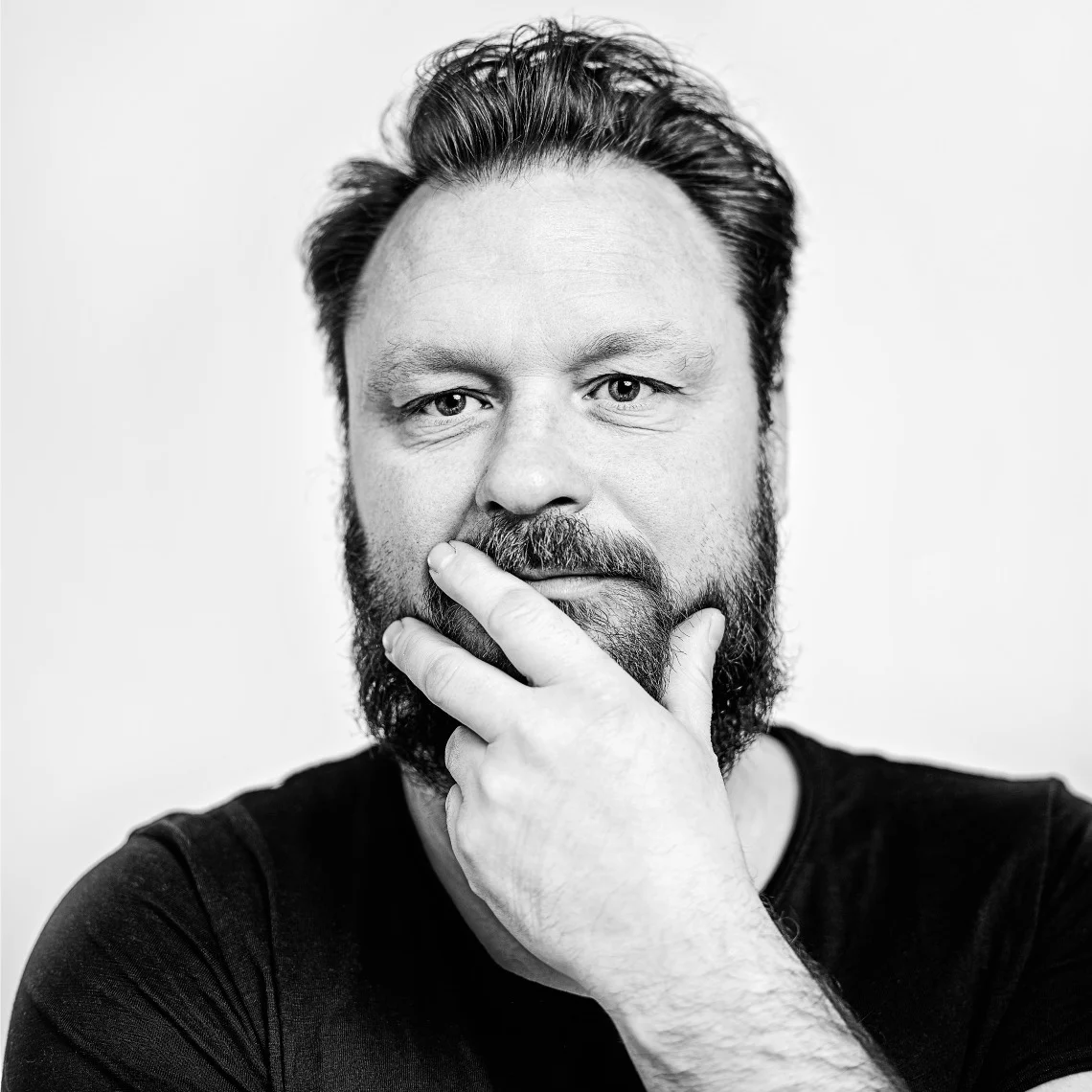 Svartvitt porträtt av en skäggig man med mörkt hår som tittar in i kameran med en hand på hakan.