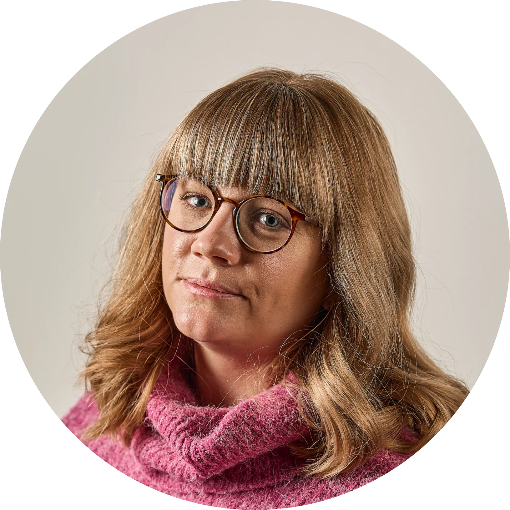 Kvinna med blont hår, lugg och glasögon, klädd i en rosa stickad tröja, tittar neutralt åt vänster.