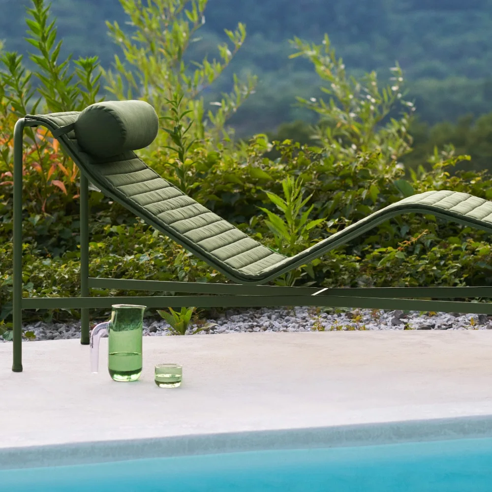 Grön solstol på en uteplats vid en pool, med en grön kanna och ett glas vatten. Frodig grönska och kullar i bakgrunden.