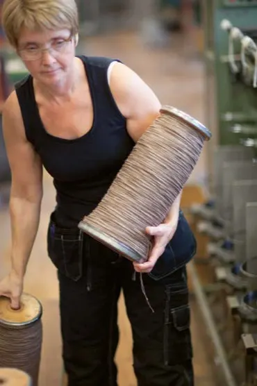En kvinna i svart linne håller en stor spole med ljusbrun tråd i en fabrik.