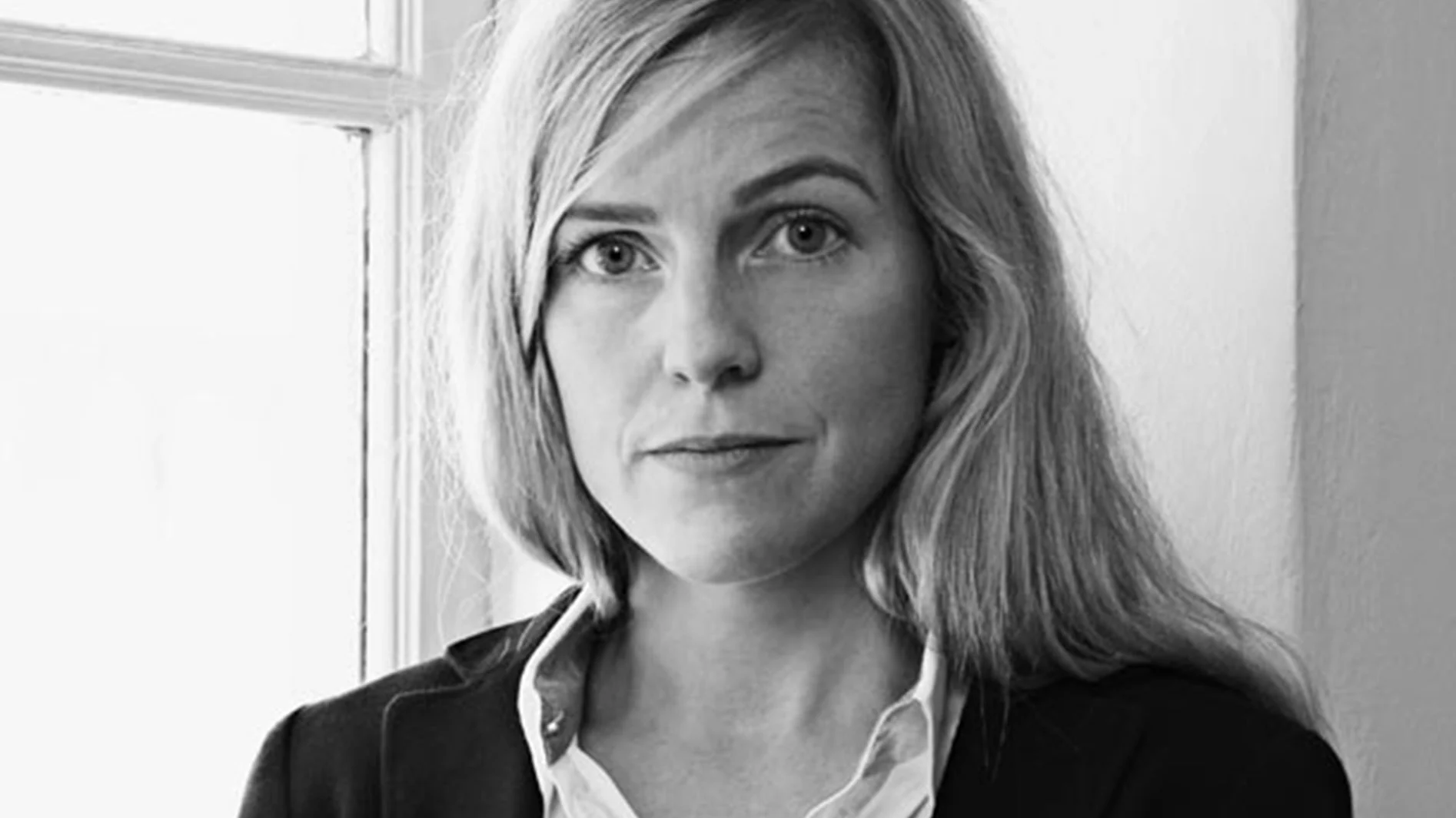 Svartvitt porträtt av en kvinna med långt blont hår, iklädd mörk jacka och ljus skjorta, som ser rakt fram.