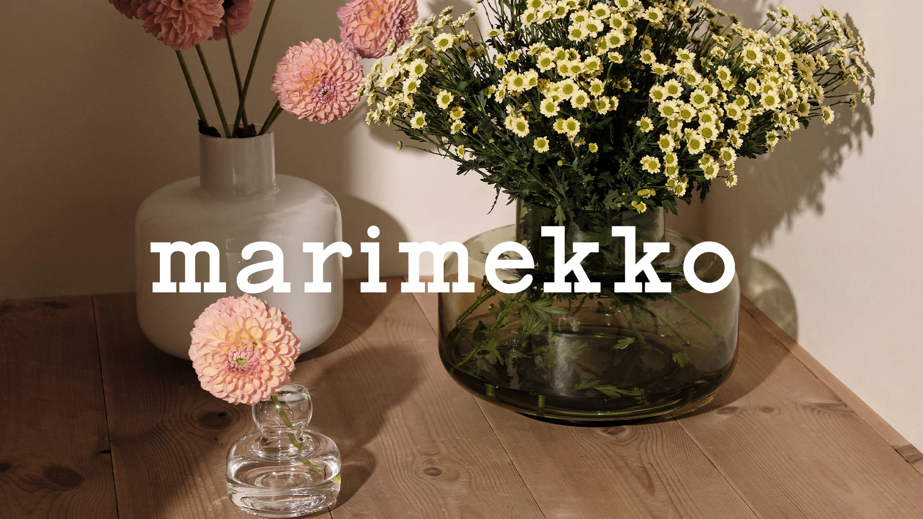 Marimekko-text över ett träbord med rosa dahlior i vita vaser och vita prästkragar i en grön vas.