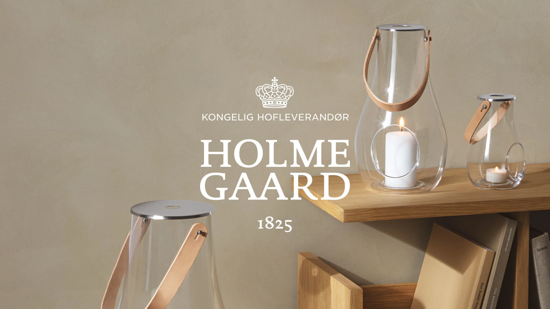 Två glaslyktor med läderhandtag i brunt på en trädhylla, en stor med tänt ljus, en liten. Holme Gaard-logga och texten '1825'.