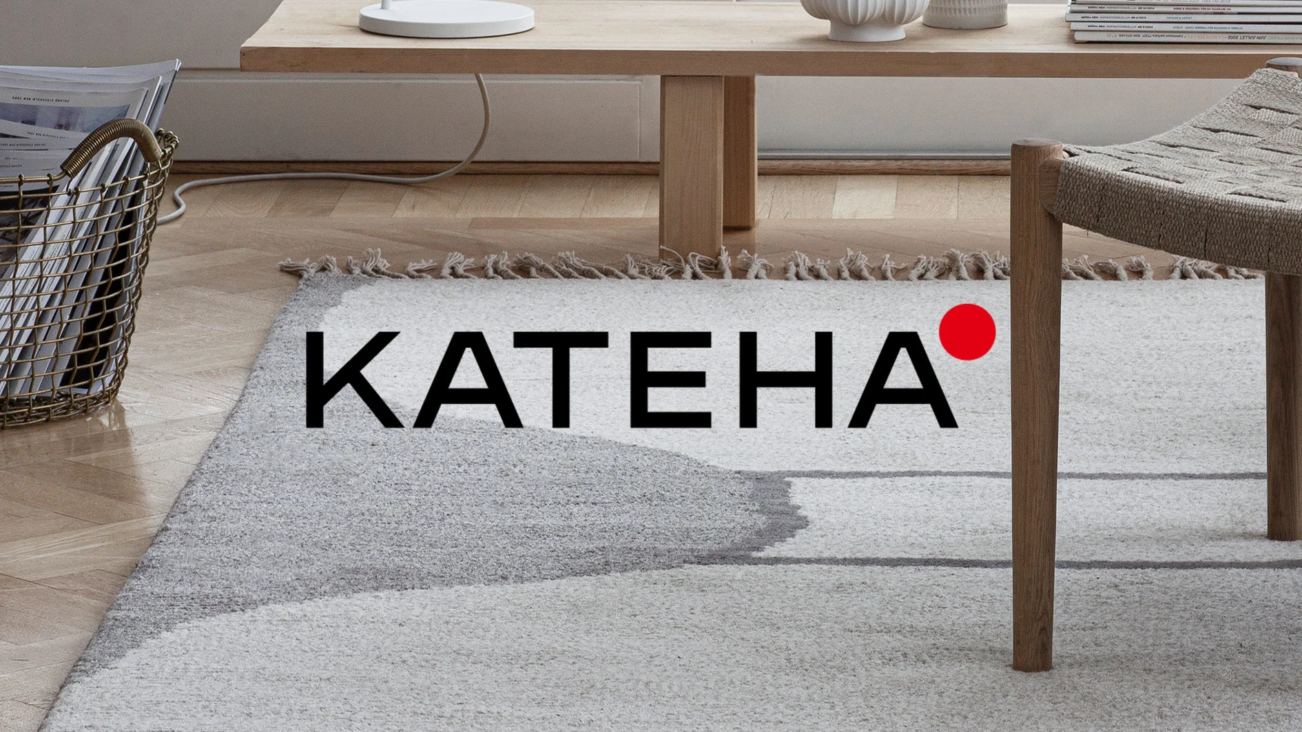 Katena-logotyp på en ljusgrå matta med abstrakt mönster, omgiven av ett bord, en stol och en tidningskorg i en modern inredning.