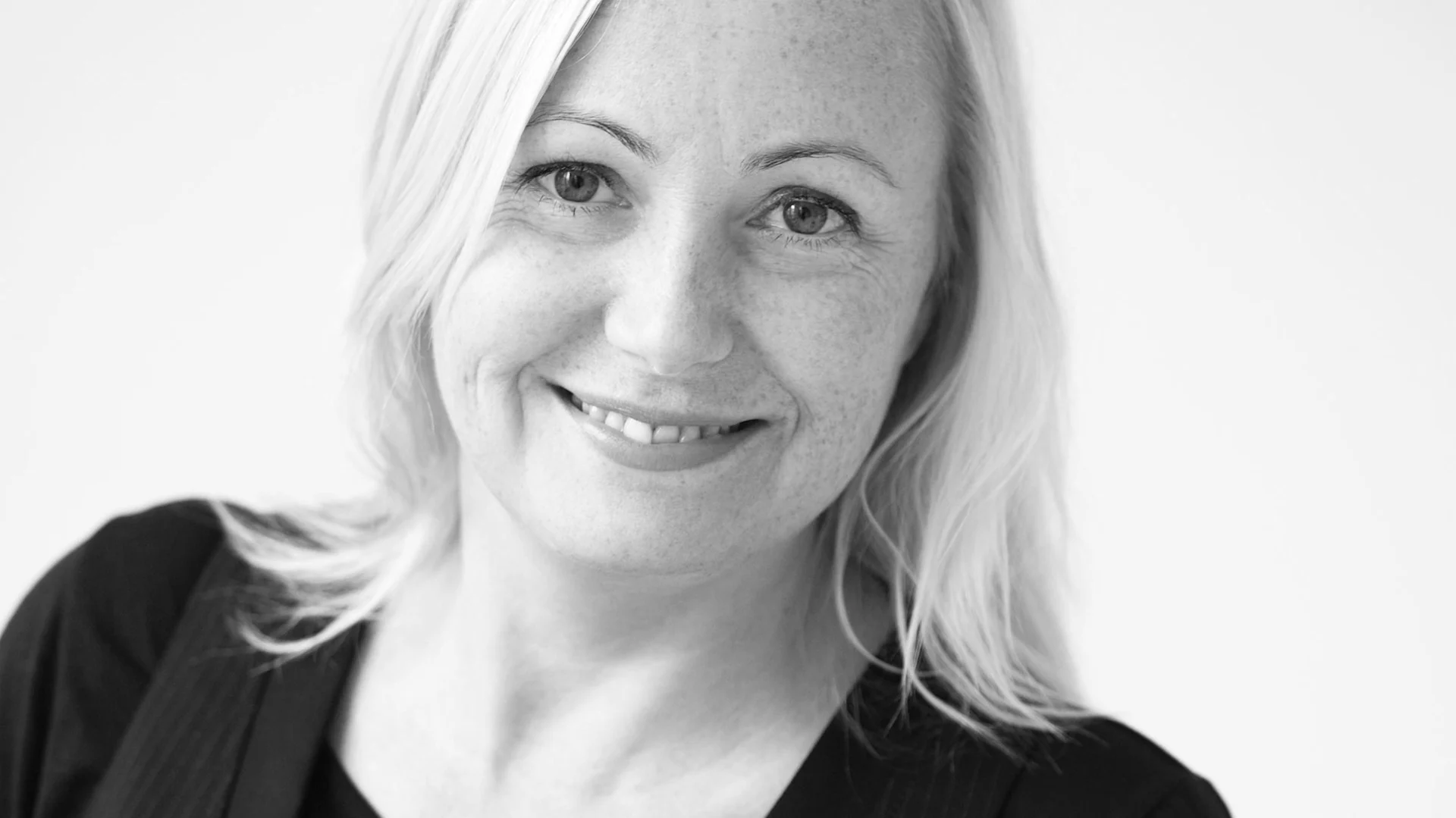 Porträtt av en leende kvinna med ljust hår och fräknar i svartvitt.