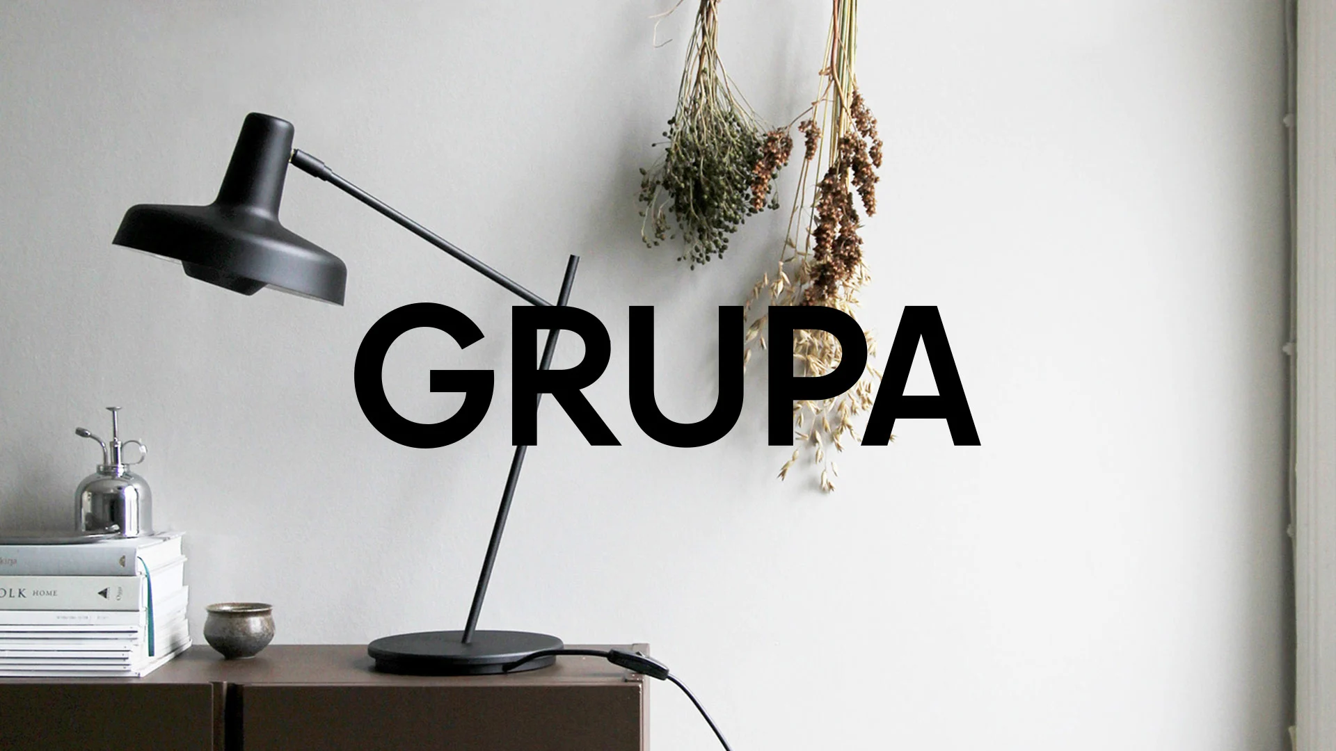 Svart skrivbordslampa, böcker och torkade växter mot en ljus bakgrund, med texten "GRUPA" över bilden.