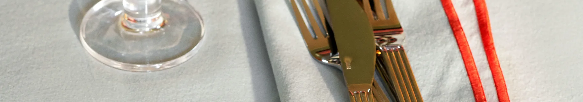 Dukning med kniv, gaffel och vinglas på ljus servett med rött band.