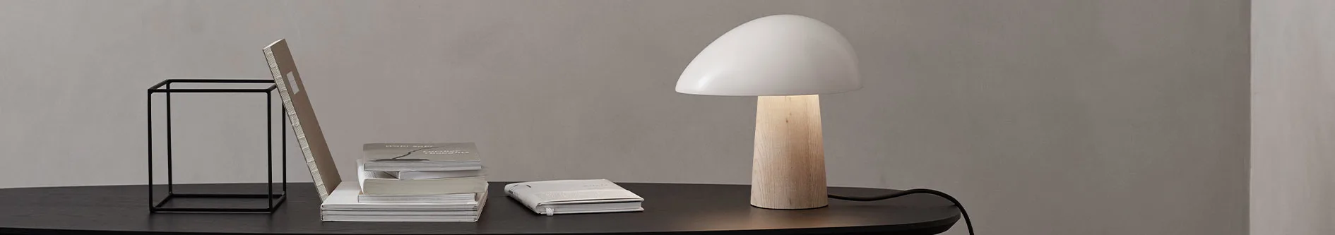 Ett mörkt träbord mot en grå vägg med en svart metallram, en bokhög och en upplyst modern lampa med träfot och vit kupolformad skärm.