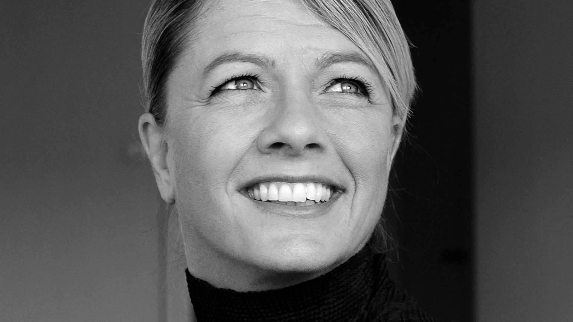 Svartvitt närbild av en leende kvinna med ljust hår som tittar upp.