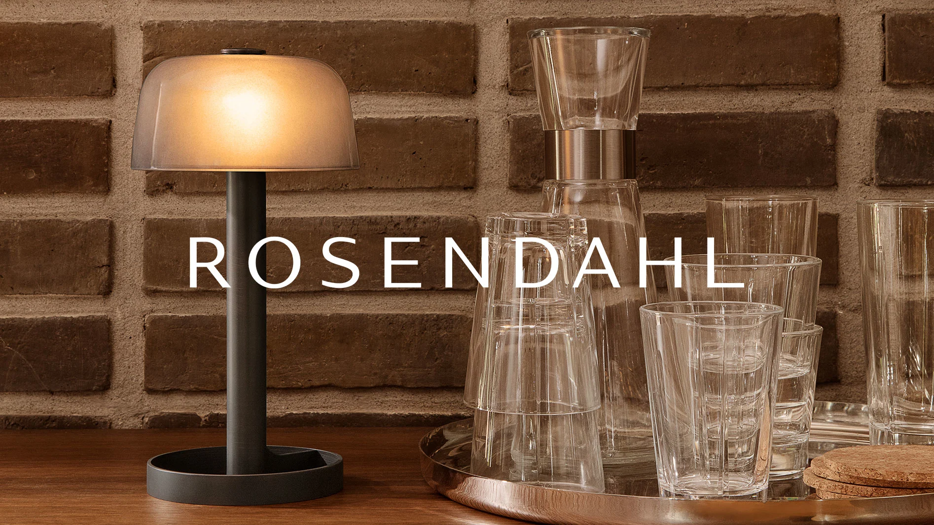 Bordslampa med lysande, klockformad skärm och ett sortiment glasvaror på en kopparfärgad bricka mot en tegelvägg.