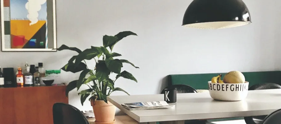 Matsalsdel med en krukväxt, ett bord med fruktskål, tidningar och en svart mugg, samt en tavla och flaskor på en skänk.