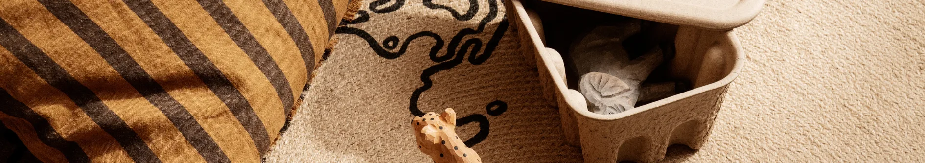 Brun- och svartrandig textil, beige vävd matta med svart mönster, fläckig träfigur och en öppen låda med en leksak.