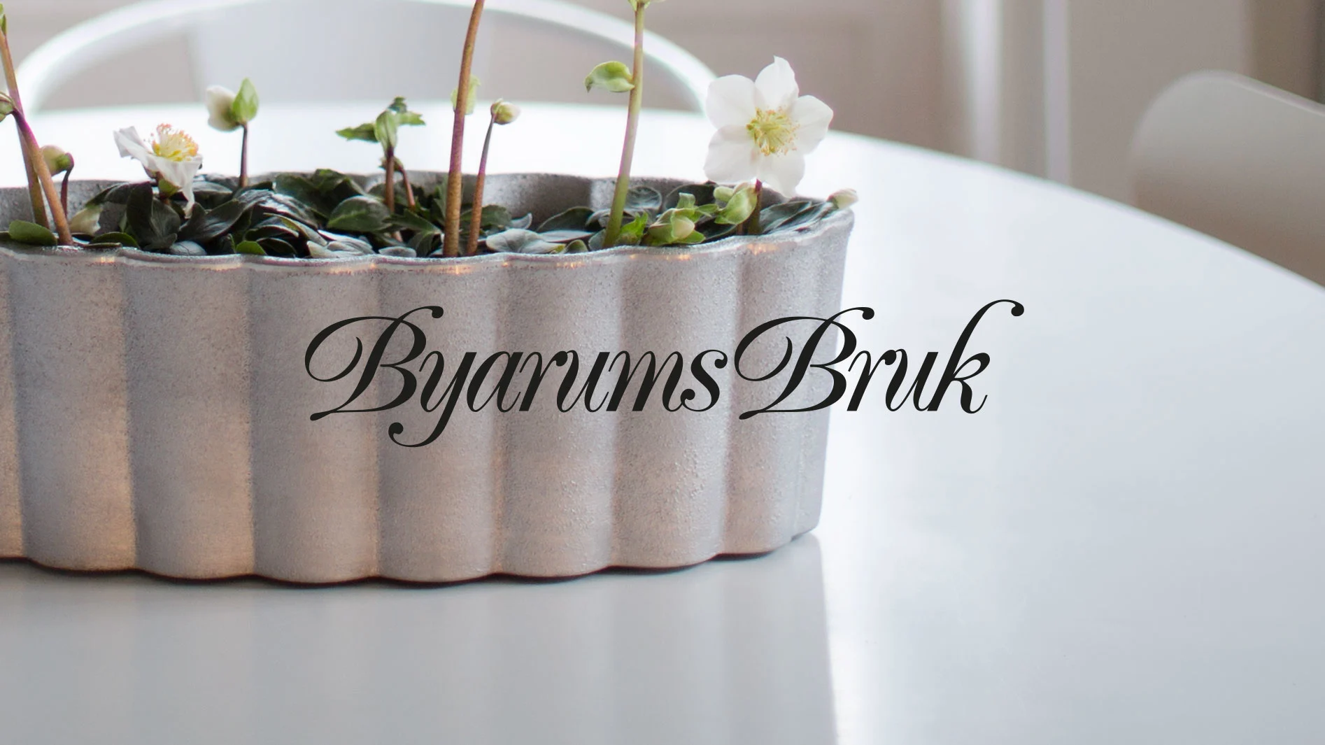 En räfflad, ljusgrå planteringslåda med vita blommor och växter, på ett vitt bord. Texten "Byarums Bruk" ligger över bilden.