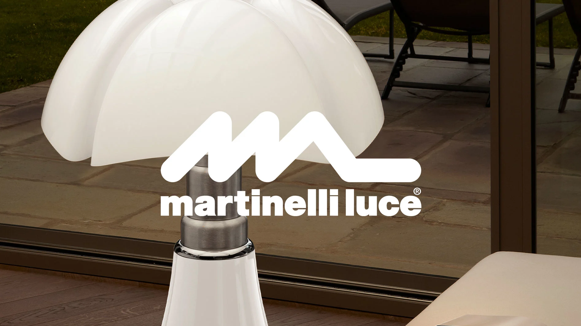 Vit svampformad bordslampa med metallbas, med Martinelli Luce-logotypen. Uteplats synlig i bakgrunden.