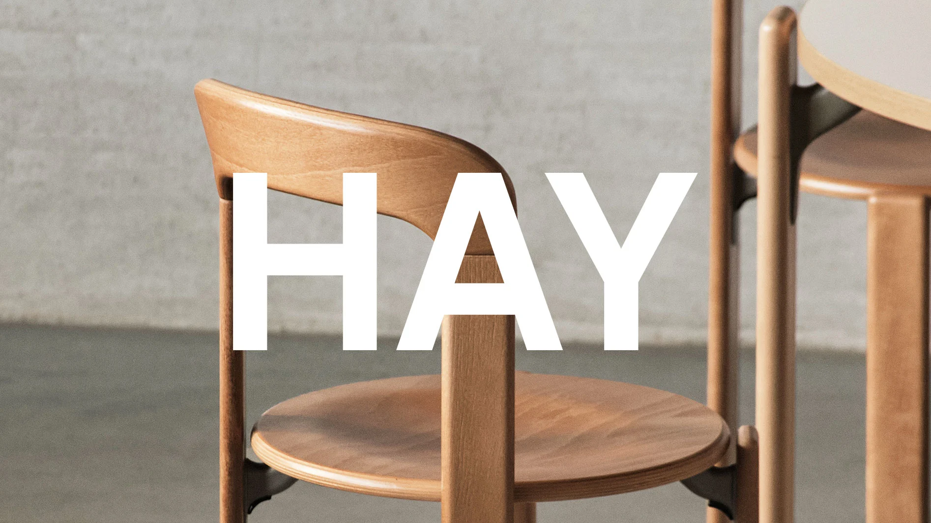 HAY-logotyp över en ljus träformad stol med böjt ryggstöd och rund sits.